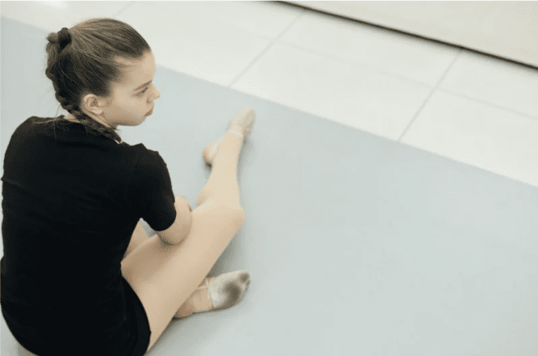 Young dancer sitting on the studio floor stretching after practice, illustrating the importance of dancer recovery and rest.